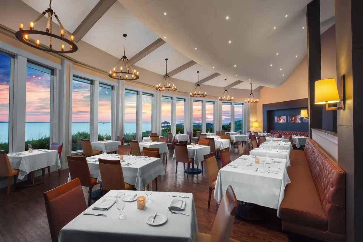 An elegant dining space with large windows framing a sunset view, chandeliers casting a warm glow, and tables set for a refined meal. The atmosphere is both sophisticated and inviting, designed for memorable dining with loved ones.