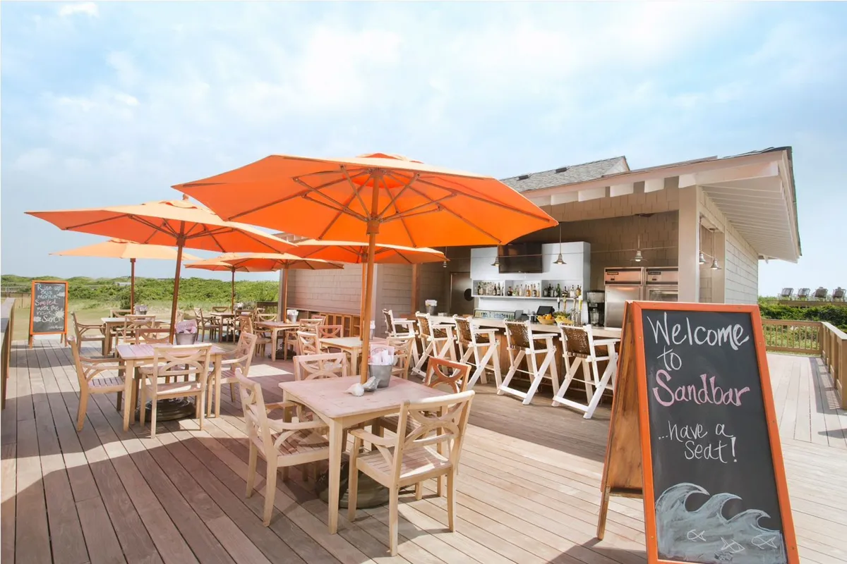 This exterior image captures a relaxed dining area with orange umbrellas, wooden tables and chairs, and a welcoming sign, all set against the backdrop of sunset views and the gentle ocean breeze. The setting is perfect for couples enjoying cocktails or an intimate meal outdoors.