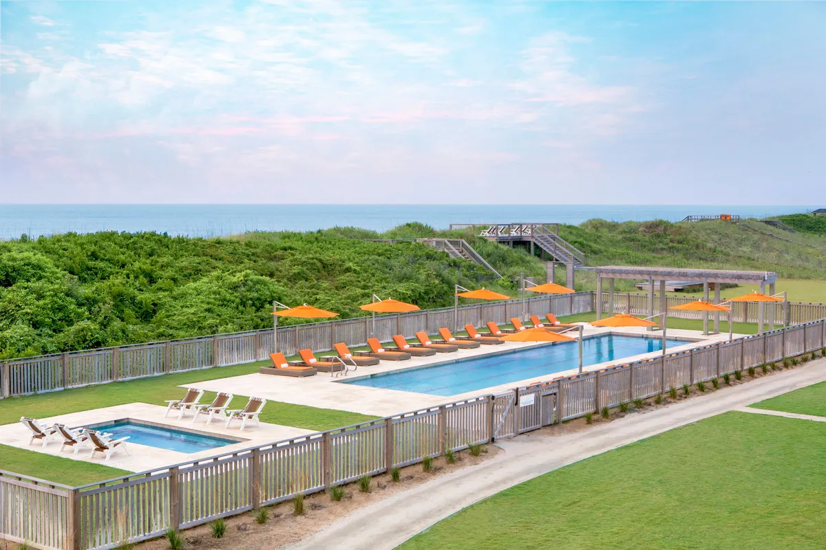 A sun-drenched outdoor pool framed by lush greenery and orange umbrellas, with lounge chairs inviting guests to relax. The pool overlooks the ocean, creating a serene atmosphere for swimming, sunbathing, or socializing. The space is alive with leisure activity and tranquil coastal views.