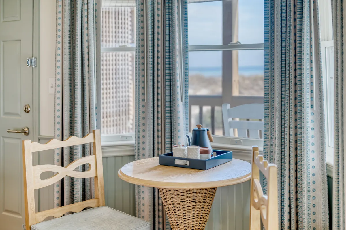 A cozy corner with a stylish wooden chair, a round table set with a teapot and cups, and patterned curtains framing a sunrise view. The inviting scene encourages slow mornings or peaceful tea breaks, emphasizing comfort and personal time.