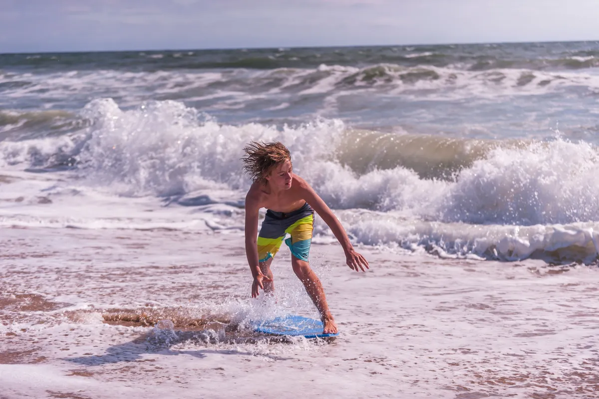 A lively beachfront scene featuring a boy riding a boogie board on the sunlit shore, with waves crashing in the background. The sandy beach stretches out, inviting family fun and adventure.