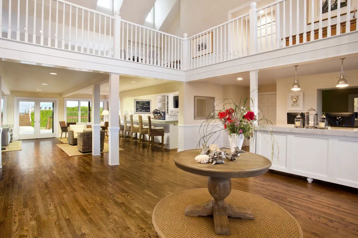 A welcoming interior scene featuring a central round table with a floral arrangement, an open kitchen in the background, and large windows leading to an outdoor area. The layout suggests an open-plan living space where dining, socializing, and family gatherings seamlessly blend together. The natural light and spaciousness highlight an environment conducive to both meals and casual lounging.