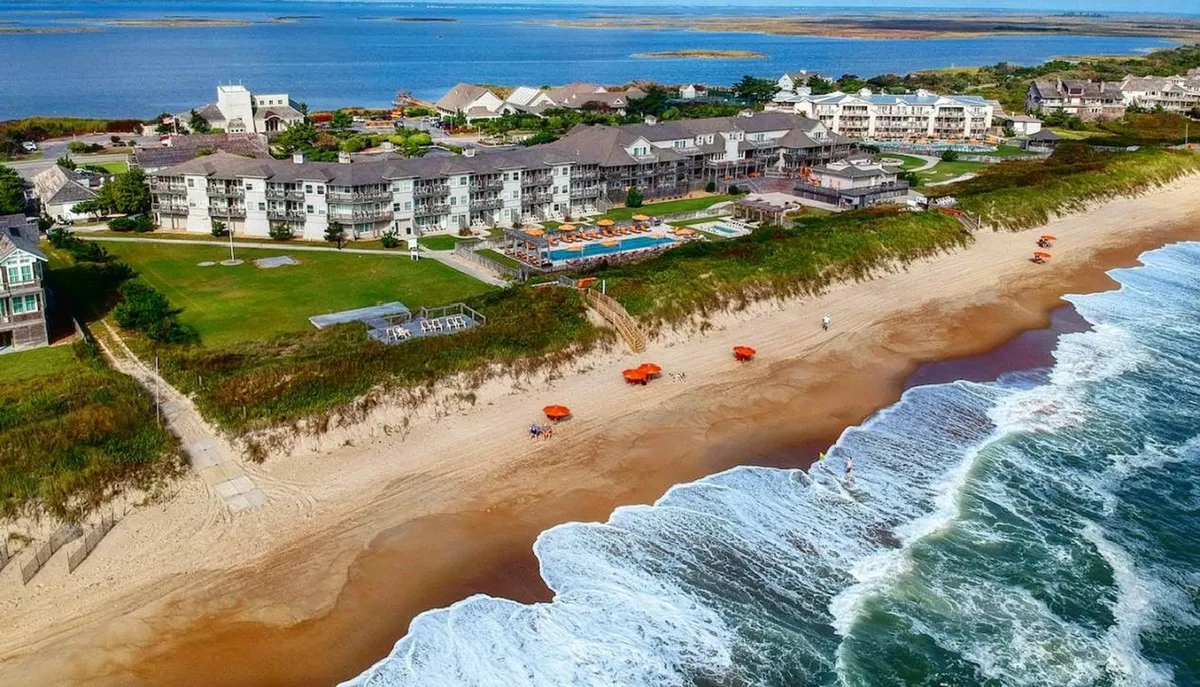 This photo captures a beachfront property with a swimming pool and colorful beach umbrellas, with the waves and shoreline just beyond. The inviting look from the balcony or pool area toward the ocean highlights the proximity to nature and the soothing sound of surf.