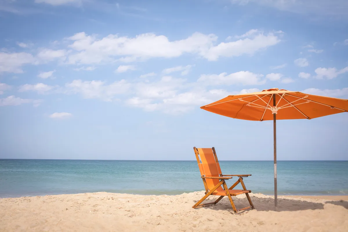 A tranquil stretch of sandy beach with a vibrant orange beach chair and umbrella set, overlooking the calm sea. The serene setting is perfect for sunbathing, relaxing, or enjoying the ocean views with loved ones.