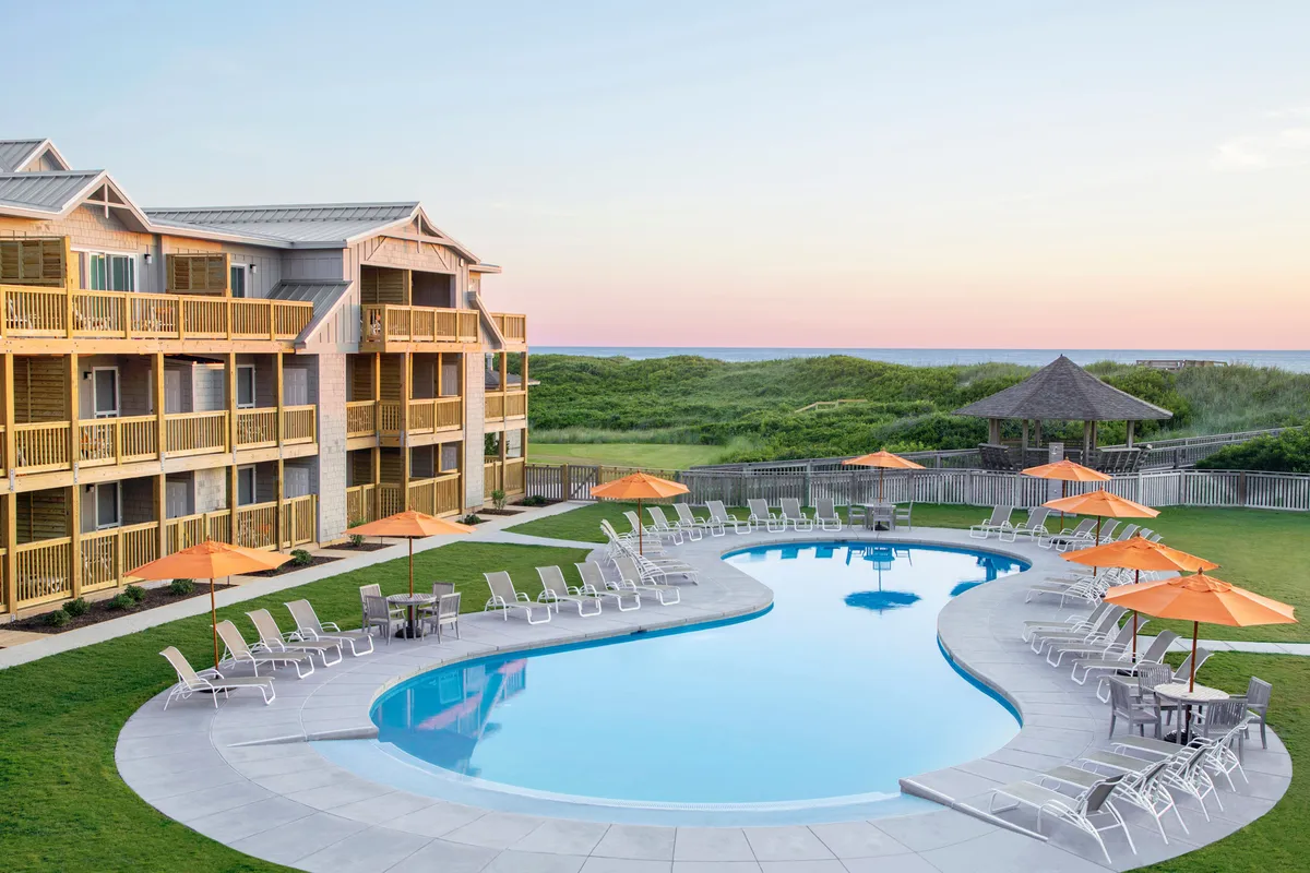 The inviting pool area at the resort, featuring a sparkling swimming pool surrounded by lounge chairs, umbrellas, and lush landscaping, with the hotel building in the backdrop. People can be seen sunbathing and socializing, embodying leisurely poolside enjoyment.
