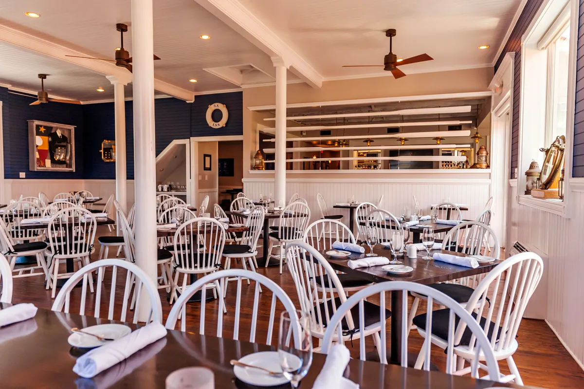 A welcoming dining room scene with tables elegantly set for guests, decorative artwork on the walls, and a stylish bar area in the background. The ambiance is lively yet sophisticated, suggesting a space ideal for shared meals and special celebrations.