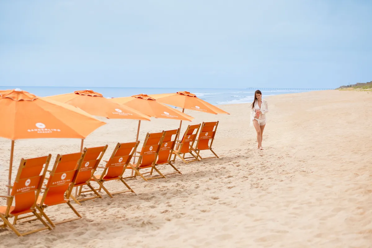 Rows of beach chairs and umbrellas line a pristine sandy beach, overlooking the tranquil ocean. A guest strolls along the shoreline, while the setup hints at seamless beach access provided by the resort, complete with all the comforts needed for a day by the sea.