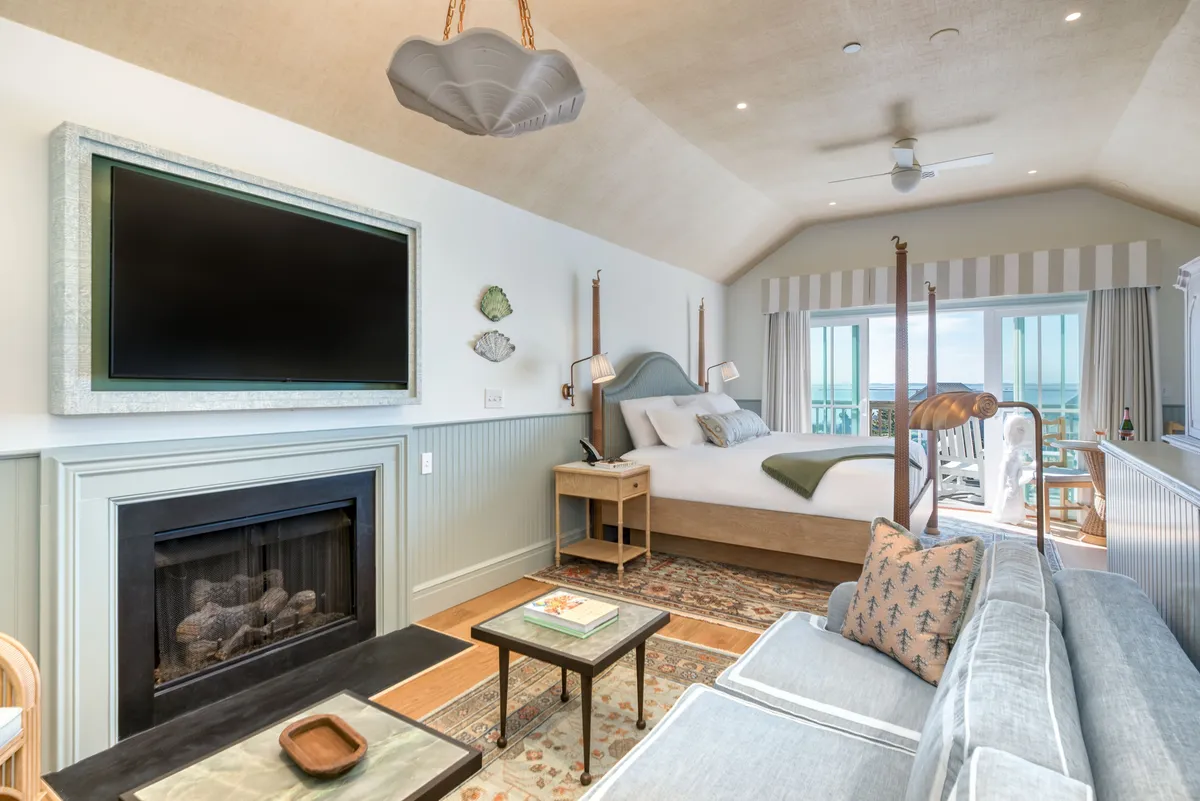 The image showcases a bright, serene studio interior featuring a luxurious king-size bed facing a cozy fireplace, with a plush sofa and coffee table nearby. Sunlight filters through large windows, offering tranquil views of the resort's natural surroundings, creating an inviting space perfect for relaxation.