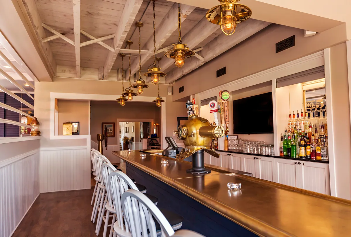 A stylish bar area featuring a brass bar top, suspended lighting fixtures, a well-stocked bottled spirits display, and comfortable wooden bar stools. The mood is welcoming, suggesting a lively spot for guests to unwind and socialize after a day of outdoor adventure.