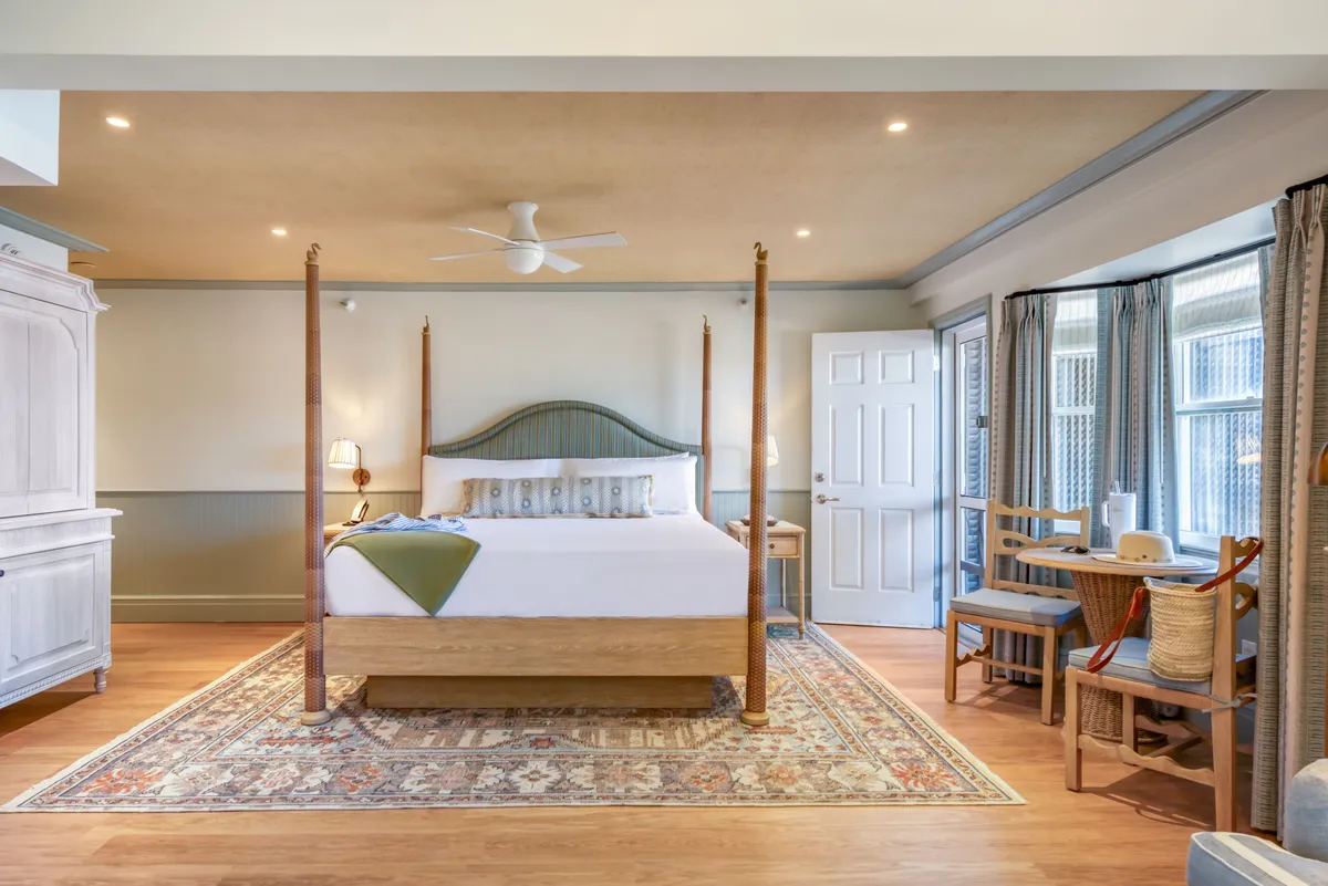 A tranquil guestroom featuring a four-poster bed with plush bedding, a decorative rug, and a window seating area bathed in soft natural light. The scene showcases a harmonious blend of comfort and understated luxury, ideal for restful sleep or morning relaxation.
