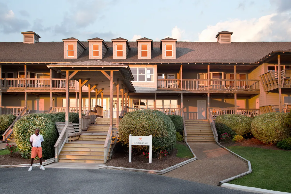 This exterior shot highlights the inviting entrance to a beach house, with steps leading up, rounded shrubs, a visible sign, and a guest standing on the porch. The scene conveys a sense of arrival and the anticipation of relaxed beachside living.