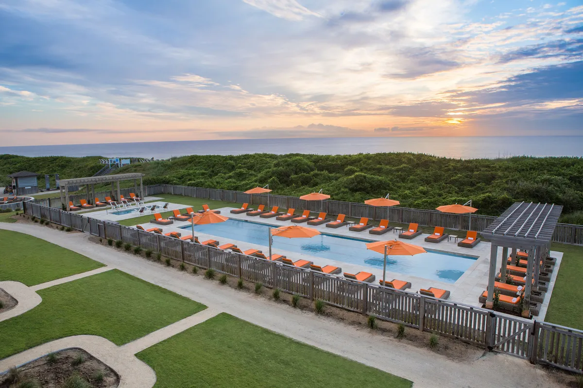 A tranquil exterior scene featuring a sparkling swimming pool lined with orange umbrellas and lounge chairs, with a beautiful sunset view in the background. The area suggests restful sunbathing, swimming, or quiet evening relaxation in a cheerful yet peaceful setting.