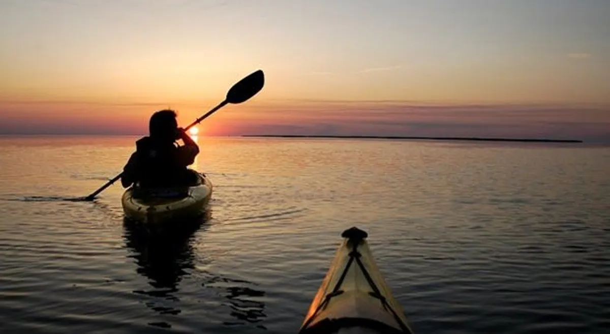A kayaker glides across calm water during sunset, with warm golden light reflecting off the gentle ripples. The expansive view of water and sky evokes a tranquil, open atmosphere, inviting guests to immerse themselves in natural beauty and relaxation.